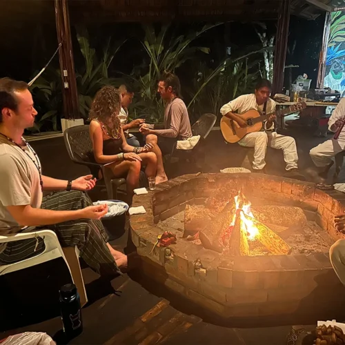 A group of ayahuasca retreat participants sitting and relaxing around a fire inside a traditional maloka in Colombia, surrounded by warm wooden walls and soft ambient light.
